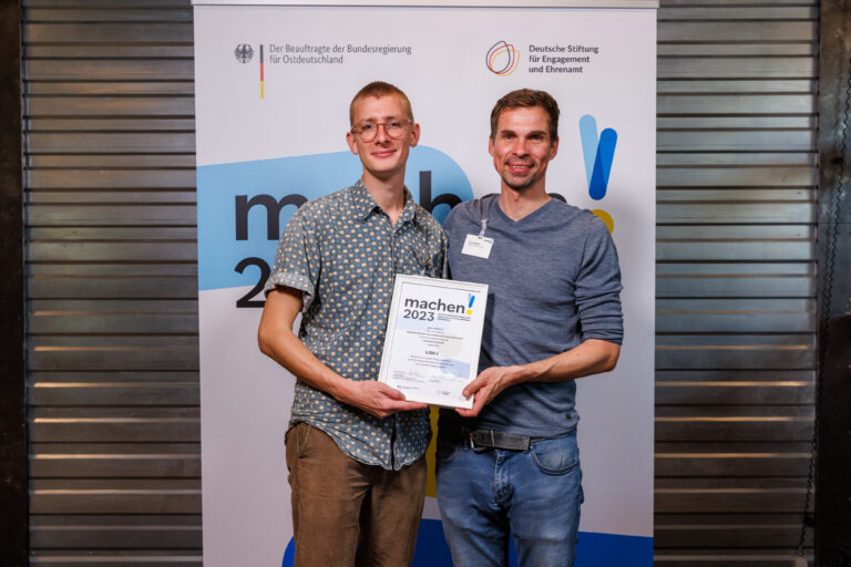 Chris und Matti vom goalsconnect e.V. stehen nebeneinander vor der Fotowand bei der Preisverleihung des Ideenwettbewerbs machen!2023 und halten ihre Urkunde in der Hand.